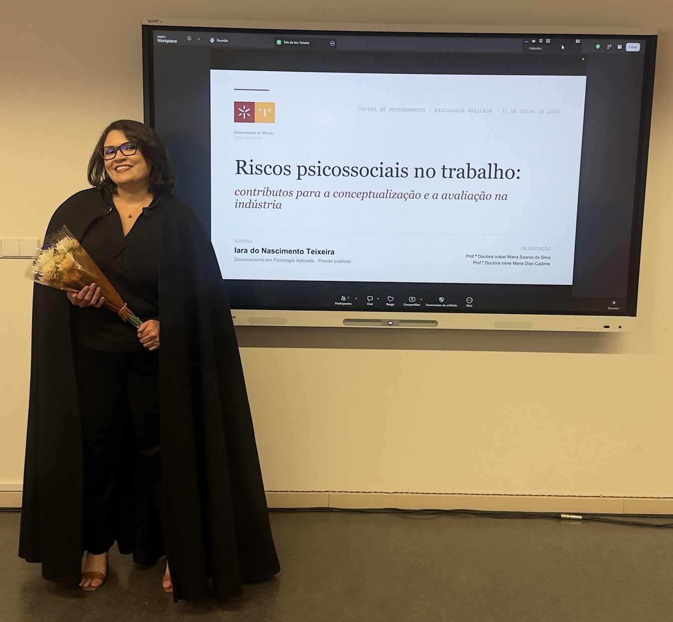 Iara Teixeira in academic dress, holding flowers, next to the opening slide of her doctoral defense at the University of Minho